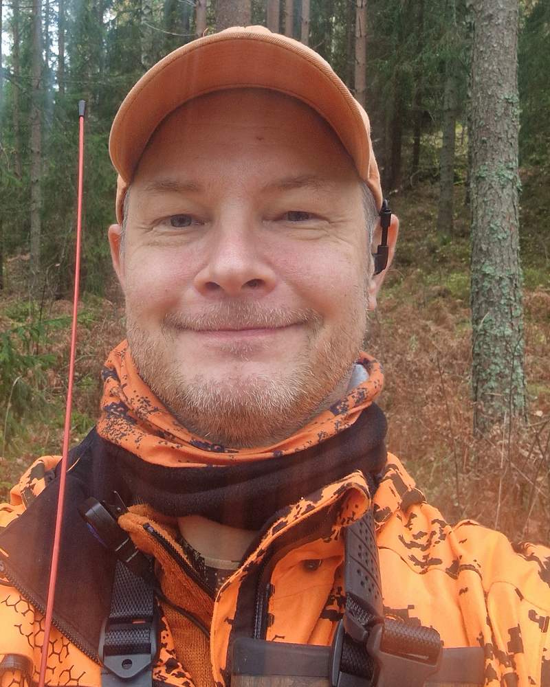 Ari Hietamäki metsässä oranssissa lippalakissa ja metsästystakissa. Kuvan reunassa näkyy radiopuhelimen antenni.